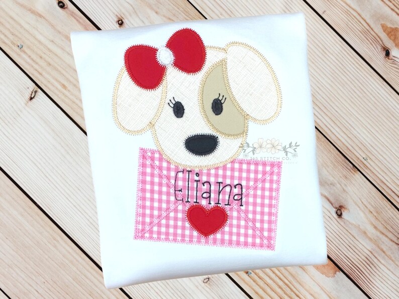 May include: White t-shirt with an embroidered applique of a dog holding a pink and white gingham envelope with a red heart and the name "Eliana" embroidered on it.