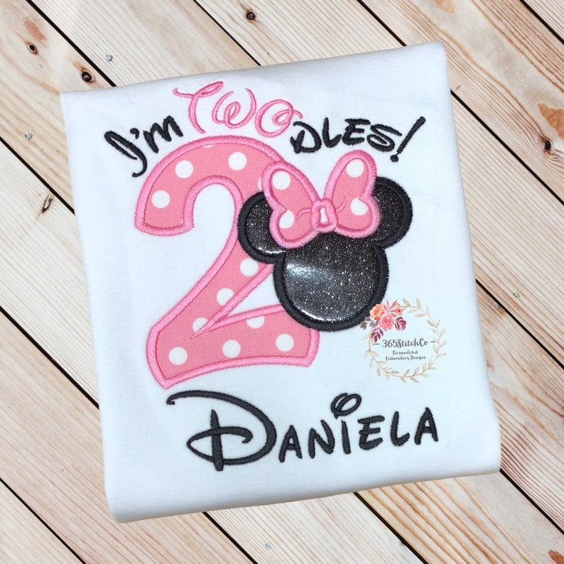 Puede incluir: Una camiseta blanca con bordados en rosa y negro. El bordado dice "I'm Two-dles!" con un n&uacute;mero 2 rosa con lunares blancos y una cabeza de Minnie Mouse negra con un lazo rosa con lunares. El nombre "Daniela" est&aacute; bordado debajo del n&uacute;mero.