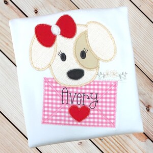 May include: A white shirt featuring an embroidered applique of a dog wearing a red bow. The dog is holding a pink and white gingham envelope with the name "Avery" written on it.