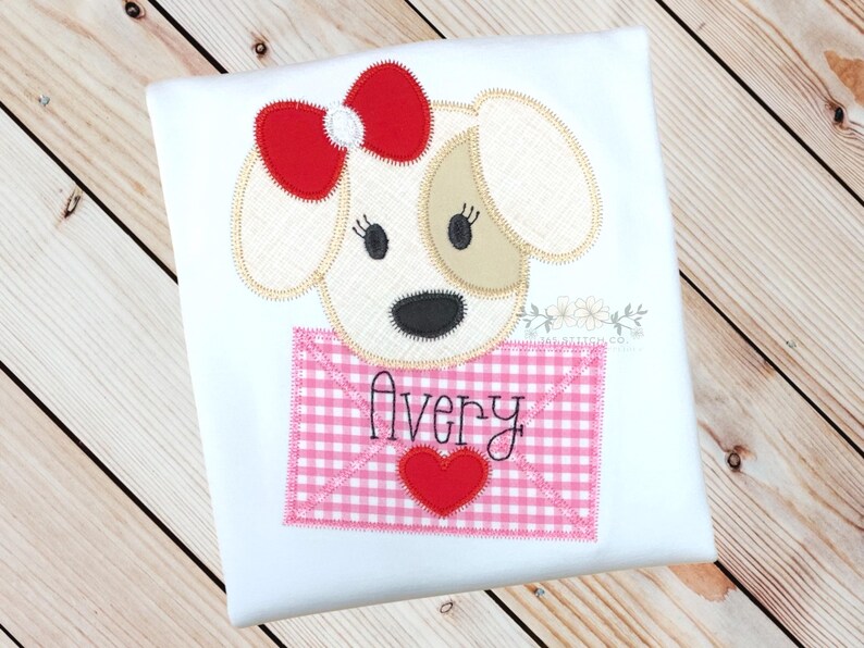 May include: A white shirt featuring an embroidered applique of a dog wearing a red bow. The dog is holding a pink and white gingham envelope with the name "Avery" written on it.