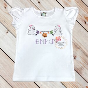 Puede incluir: Camiseta blanca con un dise&ntilde;o de fantasma y guirnalda de ma&iacute;z dulce. La camiseta tiene un nombre bordado en morado "EMMA" y un peque&ntilde;o logotipo para 365stitchco. Dise&ntilde;os de bordado personalizados.