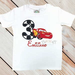 Pu&ograve; includere: T-shirt bianca con una macchina da corsa rossa e il numero 3 con un motivo a scacchi. Il nome "Emiliano" &egrave; scritto in rosso sotto l'auto. Sono presenti anche elementi floreali.