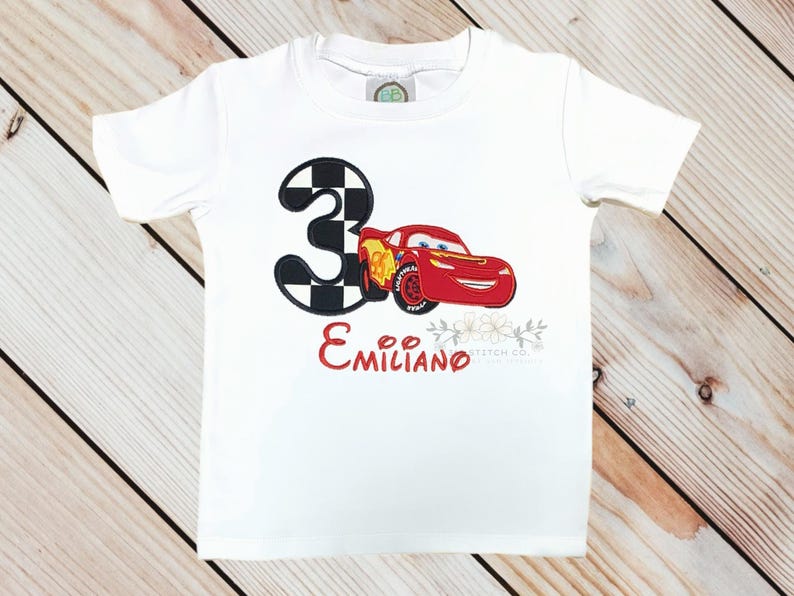 Pu&ograve; includere: T-shirt bianca con una macchina da corsa rossa e il numero 3 con un motivo a scacchi. Il nome "Emiliano" &egrave; scritto in rosso sotto l'auto. Sono presenti anche elementi floreali.