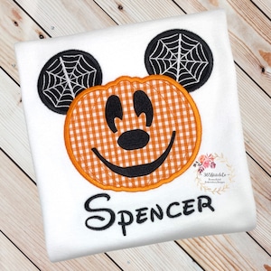 K&ouml;nnte beinhalten: Ein wei&szlig;es T-Shirt mit einem gestickten Mickey Mouse-Motiv in Form eines K&uuml;rbisses. Der K&uuml;rbis ist orange und wei&szlig; kariert, und die Ohren haben schwarze Spinnweben. Der Name "Spencer" ist unter dem Motiv gestickt.