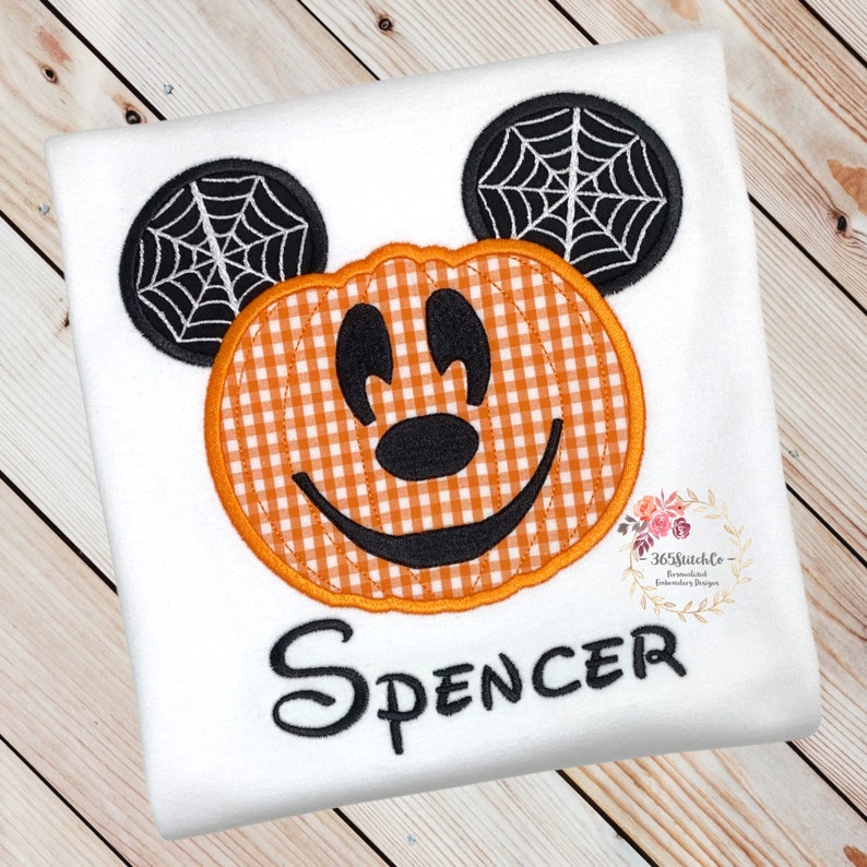K&ouml;nnte beinhalten: Ein wei&szlig;es T-Shirt mit einem gestickten Mickey Mouse-Motiv in Form eines K&uuml;rbisses. Der K&uuml;rbis ist orange und wei&szlig; kariert, und die Ohren haben schwarze Spinnweben. Der Name "Spencer" ist unter dem Motiv gestickt.