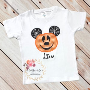 K&ouml;nnte beinhalten: Wei&szlig;es T-Shirt mit einem gestickten K&uuml;rbis-Mickey-Mouse-Motiv in Orange und Schwarz. Das Shirt tr&auml;gt auch den Namen "Liam", der unter dem Motiv gestickt ist. Das Shirt hat einen Blumengirlanden mit dem Text "365StitchCo - Personalisierte Stickdesigns" unten.