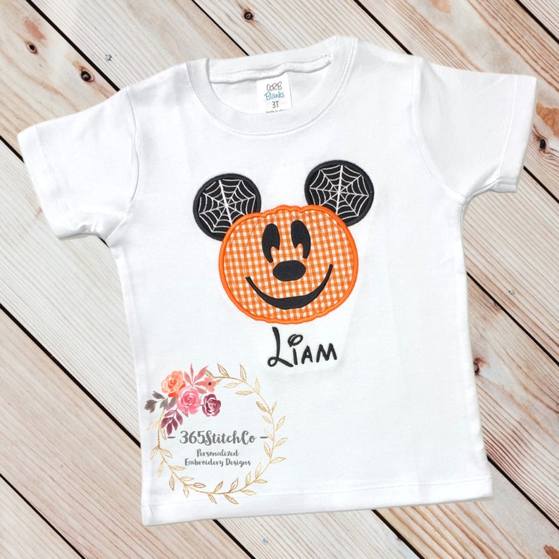K&ouml;nnte beinhalten: Wei&szlig;es T-Shirt mit einem gestickten K&uuml;rbis-Mickey-Mouse-Motiv in Orange und Schwarz. Das Shirt tr&auml;gt auch den Namen "Liam", der unter dem Motiv gestickt ist. Das Shirt hat einen Blumengirlanden mit dem Text "365StitchCo - Personalisierte Stickdesigns" unten.