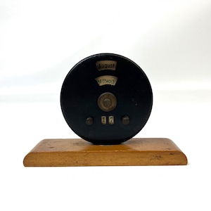 May include: A black circular desk calendar mounted on a rectangular wooden base. The calendar features windows displaying the month August, the day Mittwoch, and the date 17. A central brass-colored knob is positioned between the date windows.