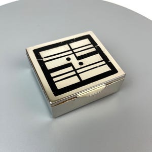 Könnte beinhalten: Kleine, rechteckige silberne Schatulle mit Scharnierdeckel. Die Oberseite der Schatulle zeigt ein schwarz-weißes geometrisches Design. Das Design besteht aus schwarzen Linien und Formen auf weißem Hintergrund. Die Schatulle wird wahrscheinlich zur Aufbewahrung kleiner Gegenstände verwendet.