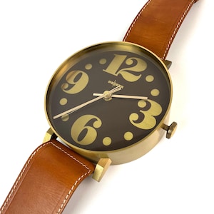 May include: A large gold-toned wall clock shaped like a wristwatch with a brown leather strap. The clock face has black background with gold numbers and a gold-toned hour and minute hand.