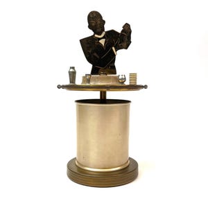 May include: A metallic bar tool set featuring a central cylindrical container on a circular base. A flat, dark silhouette of a person in a suit stands above a circular tray holding a small shaker, two cups, and a stack of circular tokens or coasters.