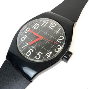 May include: A black wristwatch with a black band and a white face with black numbers and a red second hand. The watch face has a grid pattern and says "Quartz" at the bottom.