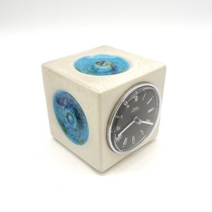 Puede incluir: Un reloj de cerámica blanca con una esfera negra y marcadores de hora con números romanos. El reloj tiene forma de cubo y tiene tres diseños circulares azules y verdes en los lados.
