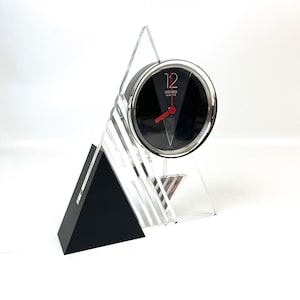 May include: A vintage Seiko Quartz desk clock with a black face, red hands, and silver trim. The clock is set in a clear acrylic and black geometric stand. The clock face displays the number 12 in a bold font.