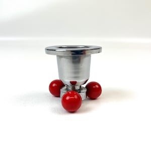 May include: A chrome-finished candle holder with a wide, flat top and a cylindrical body. Four small, bright red spheres form the base, providing stability. The design is simple and modern, suitable for various decor styles.
