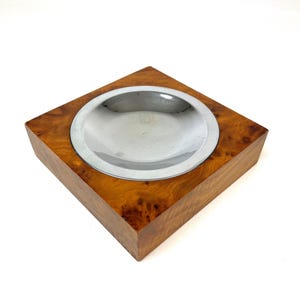 Könnte beinhalten: Quadratischer Aschenbecher aus Holz mit einem Einsatz aus poliertem Metall. Das Holz hat einen warmen Farbton mit einem natürlichen Maserungsmuster. Der Aschenbecher ist ein funktionaler und dekorativer Gegenstand.