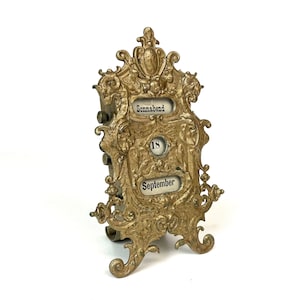 Puede incluir: Un calendario de escritorio de metal dorado con un diseño ornamentado. El calendario muestra la fecha "18" y el mes "septiembre" en letras blancas. El día de la semana es "Sonntag" en alemán.