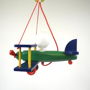 May include: A hanging pendant light fitting shaped like a biplane, featuring a green fuselage, blue wings, yellow struts, and red wheels. A single white globe light bulb is mounted on top of the plane, which is suspended by red cords.
