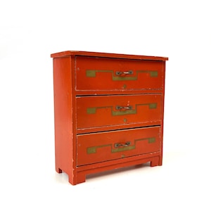 Könnte beinhalten: Eine kleine Kommode mit drei Schubladen in leuchtendem Orange. Die Schubladen sind mit messingfarbenen Beschlägen und Griffen versehen. Die Kommode steht vor einem weißen Hintergrund, der ihre Vintage-Ästhetik hervorhebt.