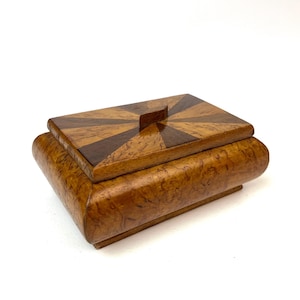 May include: A rectangular wooden box with a rounded base and a lid featuring a sunburst pattern of alternating light and dark wood veneers. The box has a polished finish and a small, centered rectangular wooden handle on the top lid.