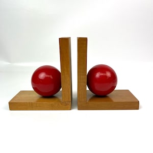 May include: A pair of wooden bookends with red ball accents. The bookends are made of light brown wood and have a simple, modern design.
