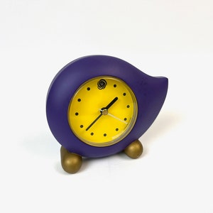 Puede incluir: Un despertador de forma abstracta de color morado con una esfera amarilla y agujas negras. El reloj tiene pies dorados.