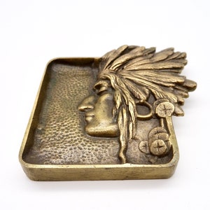 May include: A brass ashtray featuring a raised design of a Native American woman wearing a headdress. The ashtray has a textured surface.
