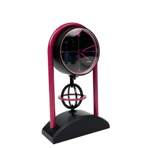 May include: A pink and black pendulum clock with a round face and a globe-shaped pendulum. The clock is made by Kundo.
