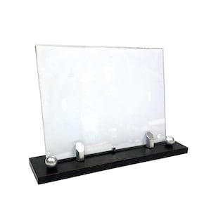 Könnte beinhalten: Ein klarer Glasbilderrahmen mit einem dunkelbraunen Holzsockel. Der Rahmen wird von zwei silberfarbenen Metallpfosten und zwei silberfarbenen Metallkugeln getragen. Der Rahmen ist für ein Foto oder Kunstwerk konzipiert.