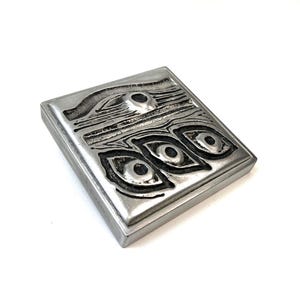 May include: A square, silver-toned metal plaque featuring a raised, textured relief design. The surface displays a series of abstract, eye-like shapes and horizontal lines with dark, recessed areas that provide contrast against the polished metal finish.