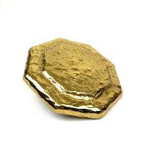 Könnte beinhalten: Ein goldfarbenes, achteckiges Objekt mit strukturierter Oberfläche. Das Objekt hat einen metallischen Glanz und scheint aus Metall zu sein. Der Artikel ist vor einem weißen Hintergrund platziert.