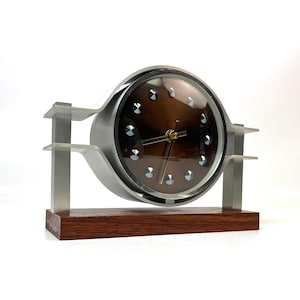 May include: A mid-century modern clock with a brown face and silver accents. The clock is set in a brushed metal frame and sits on a rectangular wooden base. The clock hands are gold. The clock is approximately 20 cm wide.