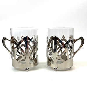 May include: Two clear glass mugs with silver-colored metal holders. The holders have an openwork design with geometric shapes and a handle. Each mug sits on three small feet.