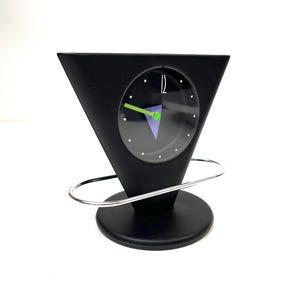 May include: A black, triangular-shaped desk clock with a circular clock face. The clock face has a black background with white dots marking the hours, and green and purple accents. The clock is supported by a silver metal ring and a black circular base.