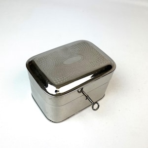 Puede incluir: Una caja rectangular de metal plateado con tapa abatible que presenta un patrón texturizado de guilloché y un cartucho ovalado central. Una llave de cuerda pequeña y delgada de metal está insertada en la parte frontal de la caja.