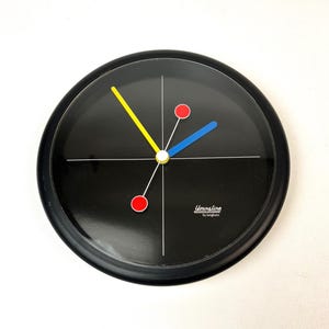 Könnte beinhalten: Eine schwarze, runde Wanduhr mit minimalistischem Design. Das Zifferblatt hat weiße Linien, einen gelben und blauen Zeiger sowie rote und gelbe Punkte. Der Text "Youngline by Junghans" ist auf dem Zifferblatt aufgedruckt.