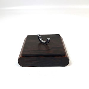 Może przedstawiać: Ciemna drewniana podstawka ekspozycyjna ze srebrnym metalowym haczykiem. Podstawka ma kwadratowy blat i fazowaną krawędź. Haczyk jest zakrzywiony i ma kwadratowy koniec po każdej stronie. Tło jest białe.
