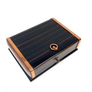 May include: A rectangular box featuring a dark, wood-grain finish with prominent black and brown vertical stripes. The lid is accented with polished copper-toned metal trim along the edges and includes a circular copper-toned ring handle.