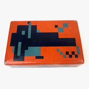 May include: Rectangular orange tin box with a pixelated design in navy blue and teal. The design appears to be a stylized dog. The box has a slightly distressed appearance.
