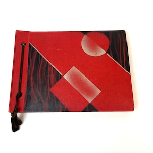 May include: A red and black art deco style photo album with a geometric design. The cover features a red background with a white circle and rectangle, and black diagonal lines. A black tassel hangs from the spine.