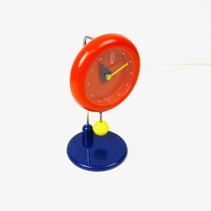 Puede incluir: Un reloj rojo con una aguja de segundos amarilla y una aguja de horas azul. El reloj está en un soporte azul con una bola amarilla.