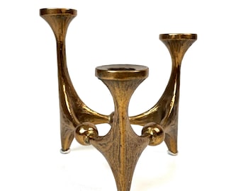 May include: A gold-colored, three-armed candle holder with a textured surface. The candle holder has a tripod base with small, round feet. The arms curve upwards, each with a circular cup to hold a candle. The design is mid-century modern.
