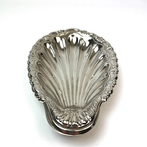 Può includere: Un piatto in metallo color argento a forma di conchiglia con una superficie lucida e riflettente. La ciotola presenta profonde scanalature radiali e un bordo decorativo smerlato con intricati dettagli in rilievo lungo tutto il perimetro.