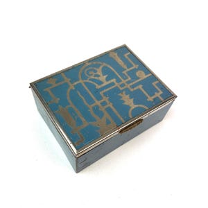 May include: A rectangular, blue box with a silver trim and a silver geometric design on the lid. The box is closed and sits on a white surface. The box is likely used for storing small items.