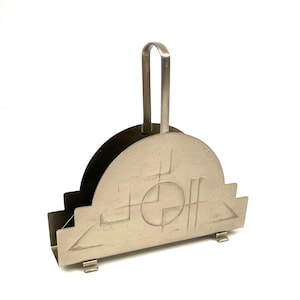 May include: A silver-toned metal napkin holder with a semi-circular top and a handle. The front features an etched geometric design. The holder has a stepped base and small feet, suitable for holding napkins or letters.