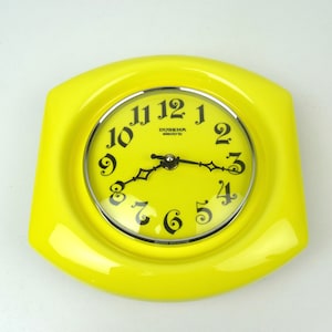 Könnte beinhalten: Eine gelbe Wanduhr mit schwarzen Zahlen und Zeigern. Das Zifferblatt hat einen silbernen Rand und trägt die Aufschrift "DUgena electric".