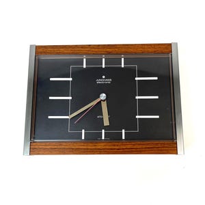May include: Rectangular Junghans electronic clock with a dark face, silver hands, and a red second hand. The clock has a brown wooden frame with a silver metal border. The clock face has white lines and the text "Junghans electronic" and "ATO T".