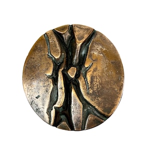 May include: A circular, bronze-toned metal item featuring a textured surface and a dark, abstract pattern. The design comprises interconnected, jagged lines and shapes. The object has a slightly uneven form and is set against a white backdrop.