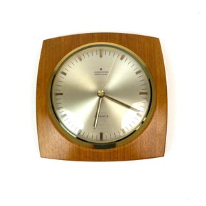 May include: A square, wooden wall clock with a gold-rimmed face. The clock face is white with black markings and gold hands. The clock face reads "JUNGHANS  Made in Germany" and "D-MAT 5".