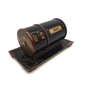 May include: A black and brass desktop calendar with a cylindrical design. The calendar displays the date "14" and the month "MAI".